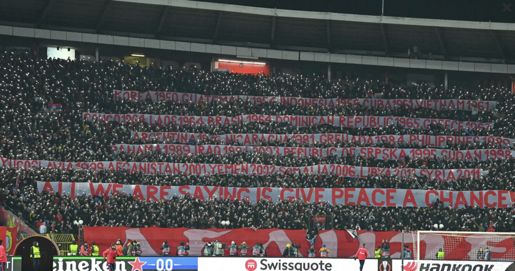 A packed crowd of Serbian sports fans hold up hundreds of glowing cell phone lights and lift aloft enormous red and white banners that spell out the United States' military interventions. The final banner reads "All we are saying is give peace a chance!"